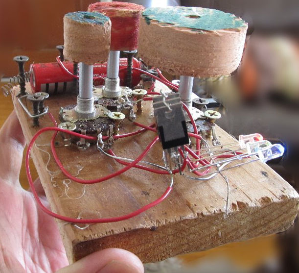 A small wooden board. Above it is circuitry, a battery, three raised wooden knobs colored green, red, and blue, and three red, green, and blue LEDs on the right.